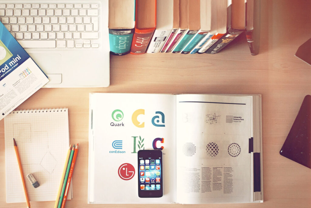 A table with a computer, iphone, design book and notepad and pencil