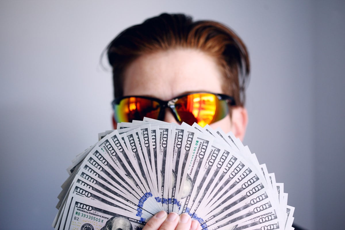 Person with sunglasses and a fan of money in front of the nose and mouth